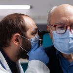 Doctor examining a senior man's ear with an otoscope during a medical checkup, both wearing face masks.